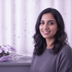Headshot of a smiling South Asian woman, late 20s, with natural makeup and casual hair, against a soft lavender and white bac